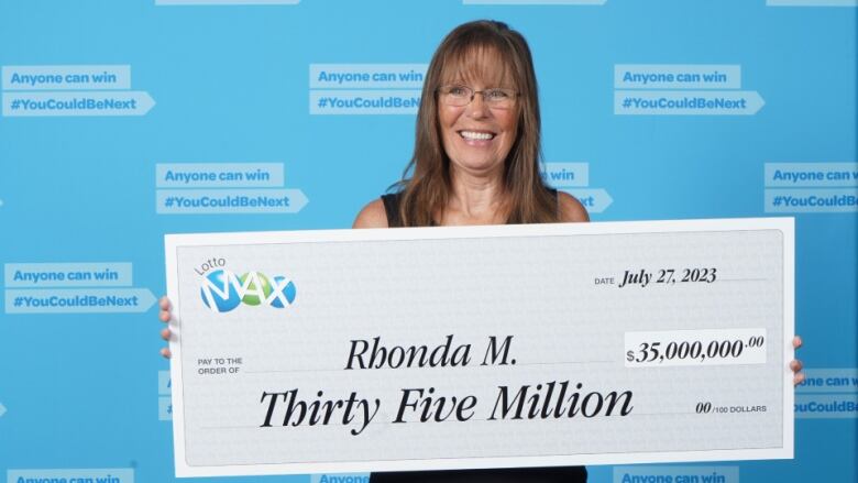 A woman poses in front of a blue background filled with slogans that all say, 'Anyone can win,' grinning and holding a giant cheque that says: Thirty-five million.