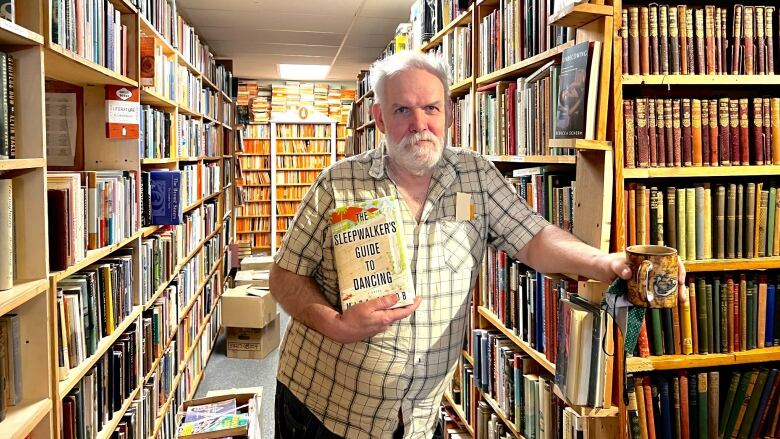 A man with white hair and a beard holds a book inside a crowded bookstore. He stands in between two shelves stacked with books. 