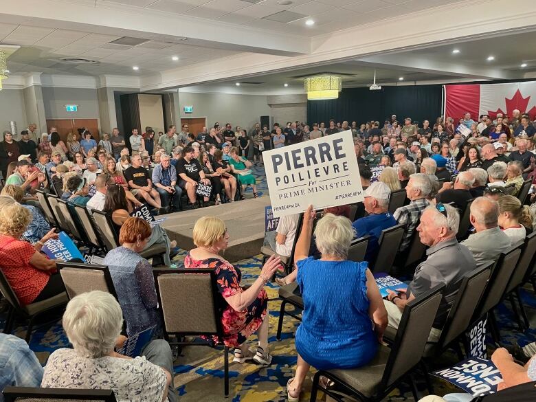 A large group of people sitting in a big room. One person is holding a sign that says Pierre Poilievre.