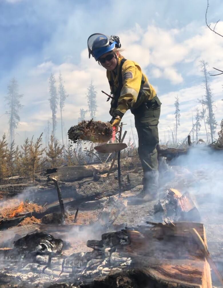 A person in firefighting gear holds up a shovelful of smouldering earth.