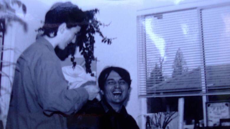 In a blue-tinged, black and white photograph, a man in a dark collared shirt is seen laughing, facing the camera. Standing next to him is a woman in an oversized shirt, smiling at him. 