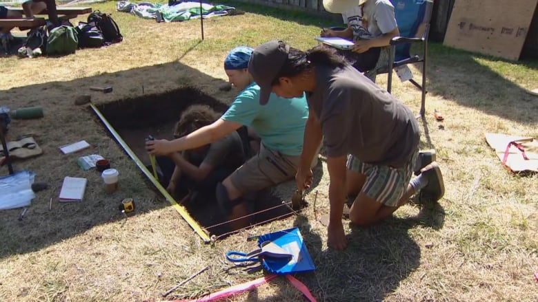 Two people are pictured in a small pit, and one squatting above it, where they are excavating for archaeological purposes