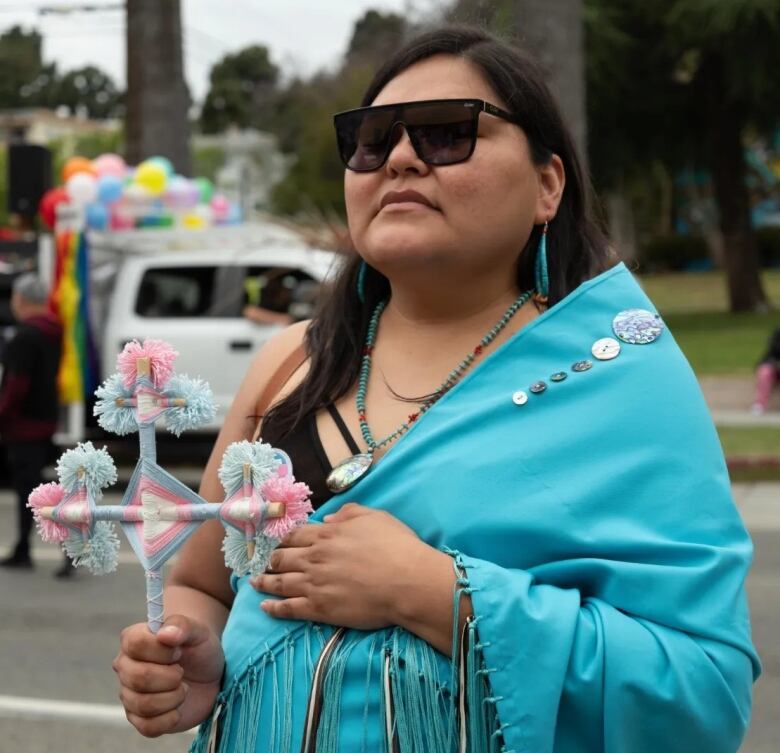 A transgender woman from the Navajo Nation, holds a cross and is wrapped in a blue blanket at a public event 