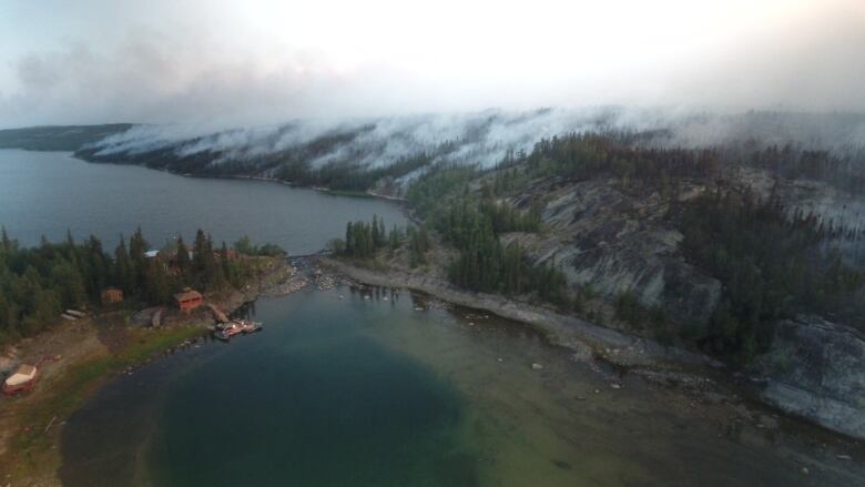 A lake with a lodge and smoke from wildfires in the woods near it. 