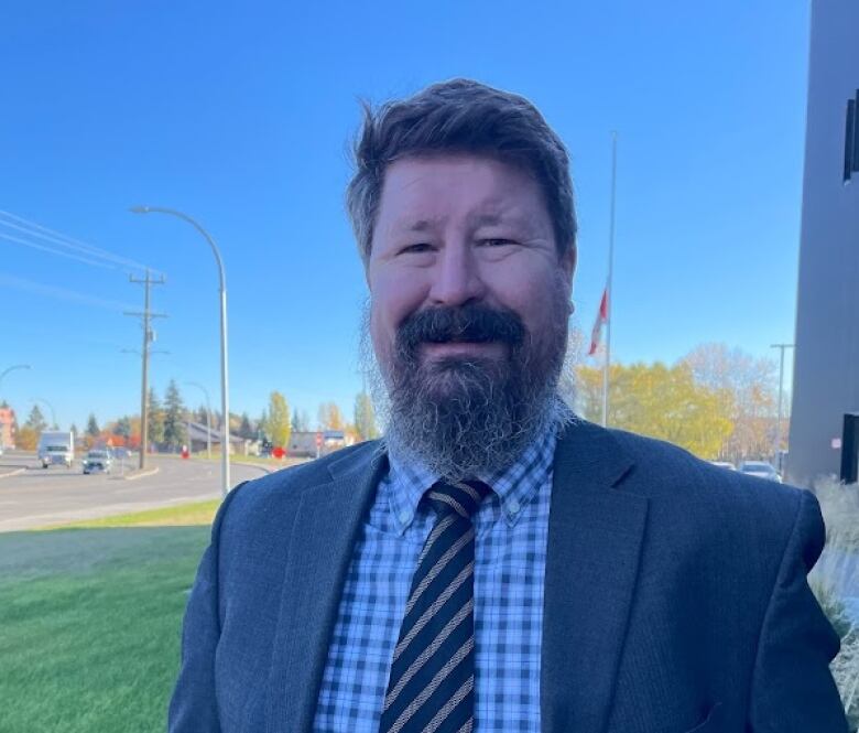 A man with a beard stands in a suit, tie and shirt, standing in front of a road.