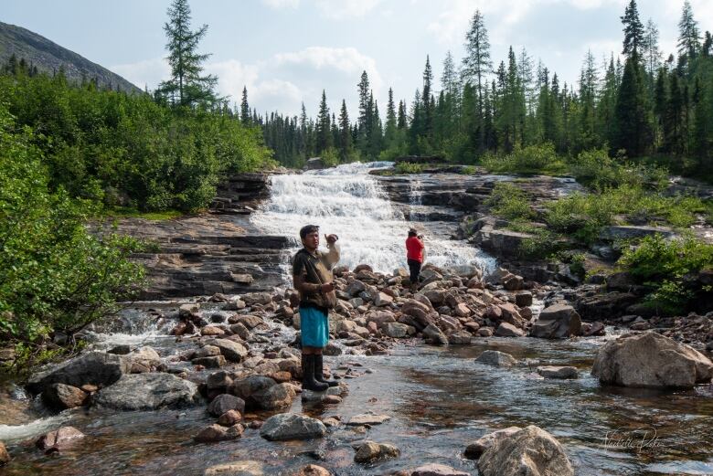 Two boys stand on rocks in a river. 