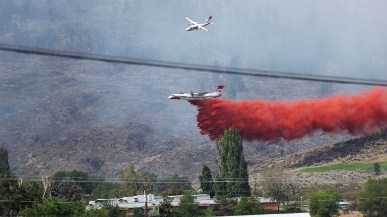 An air tanker drops bright red fire retardant on a burning hillside. Grey smoke fills the air.