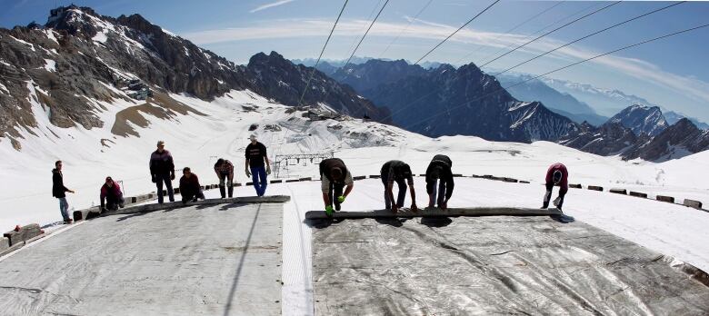 Ten people unroll large plastic sheets in a snowy mountain range that stretches into the distance.