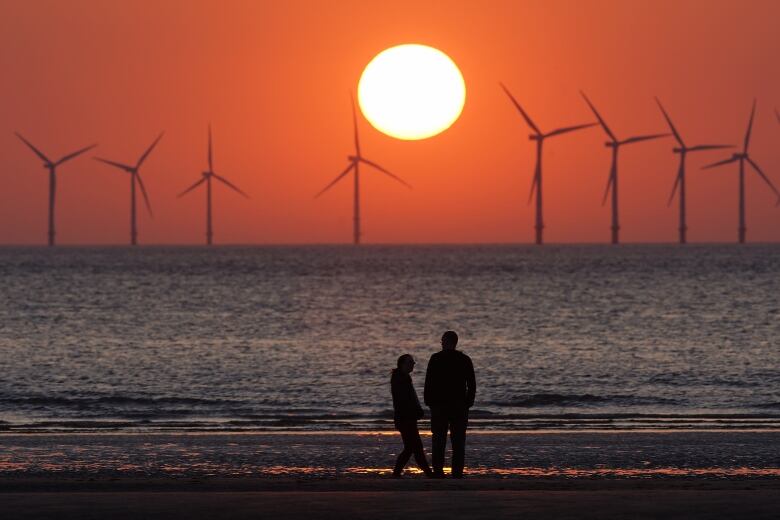 Two people are silhouetted as the sun, looking large, sets in orange sky. Eight windmills are also silhouetted in the distance, amid a calm, rippling body of water.