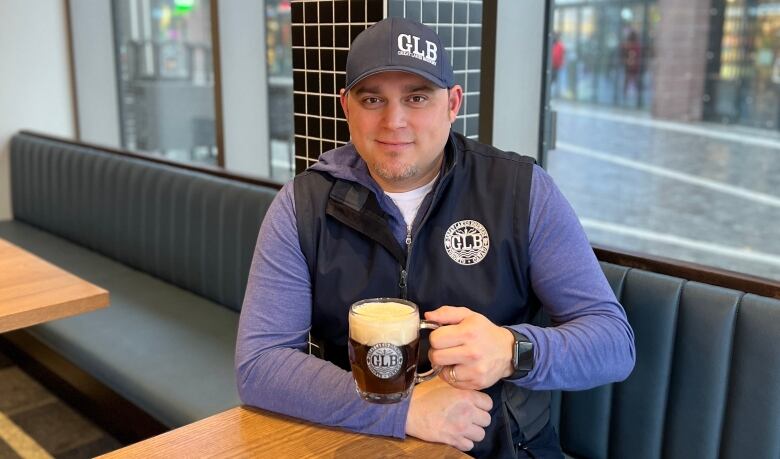 Burtch is shown sitting at a table and holding a pint glass full of beer.