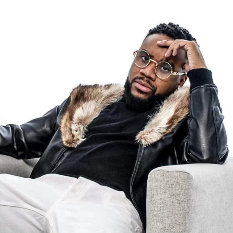 Publicity shot of Black man wearing glasses, he is seated with one hand touching his forehead.