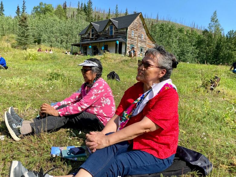 Two women sit on a grassy hillside in the sunshine, with a log building visible on a hill behind them.