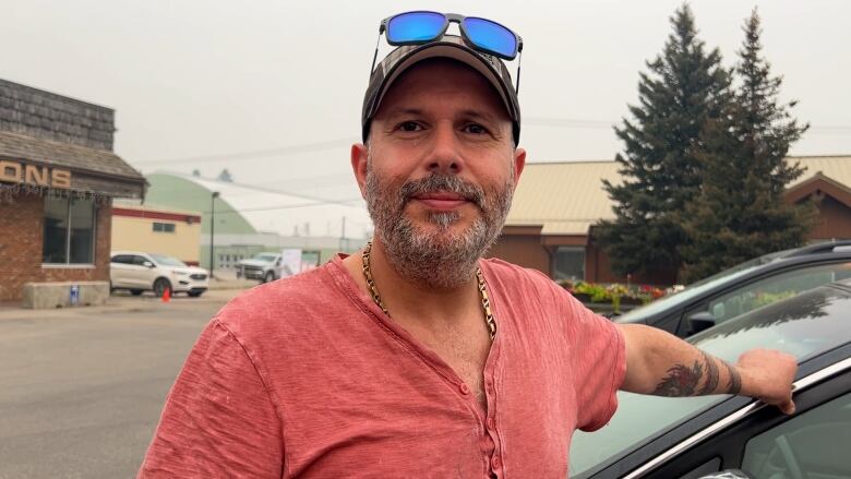 A man stands on a town street, leaning with his arm on a vehicle.