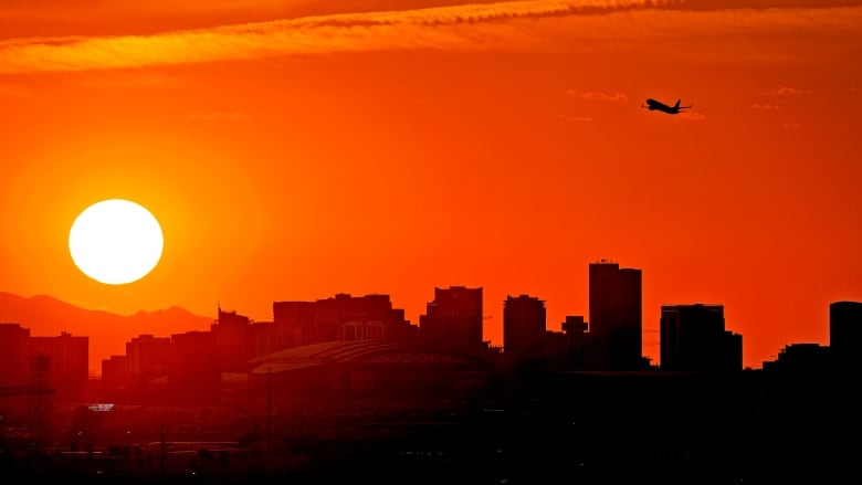 A plane in the air and buildings are silhouetted as the sun sets in the background.