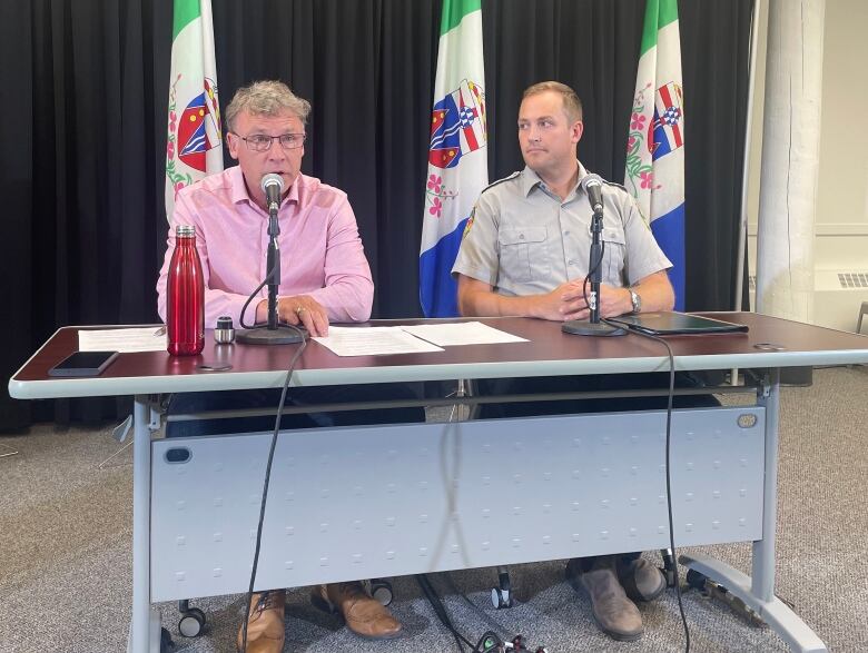 Two men sit at a table behind microphones, with a row of flags standing behind them.