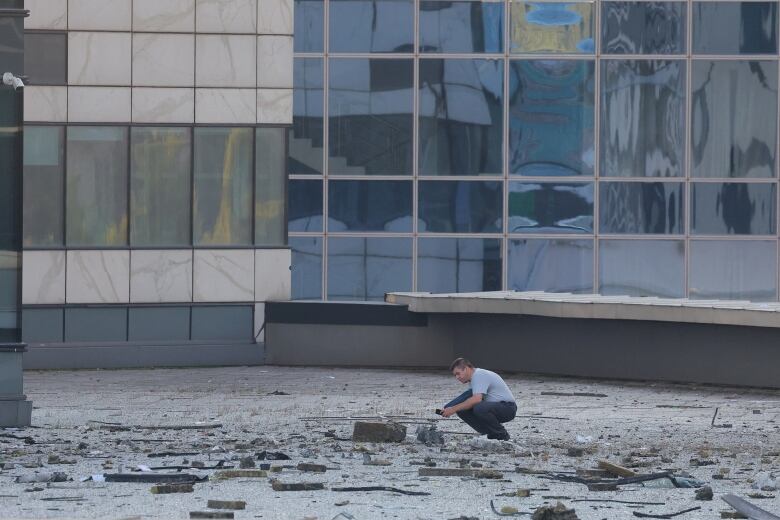 A man is shown crouching on the sidewalk with large chunks of debris and glass strewn all around him.