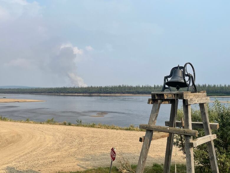 Smoke billows from a forest in the distance, across a river beside a dirt road.