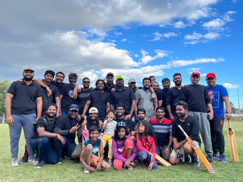 A group of men is seen posing for a photograph with kids on a cricket pitch.