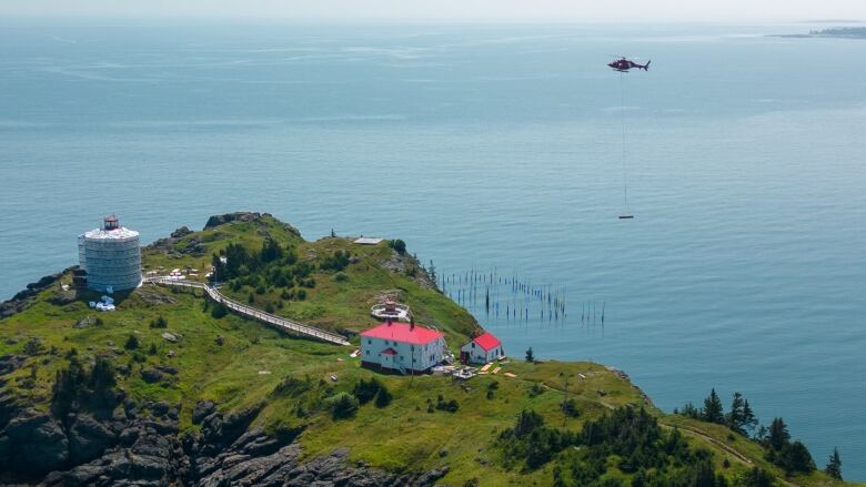 Bright and lush green island in the middle of calm blue water. A large white house with a red roof is in the middle of the island. Near the peninsula is a white lighthouse boarded up with tarp during construction. 