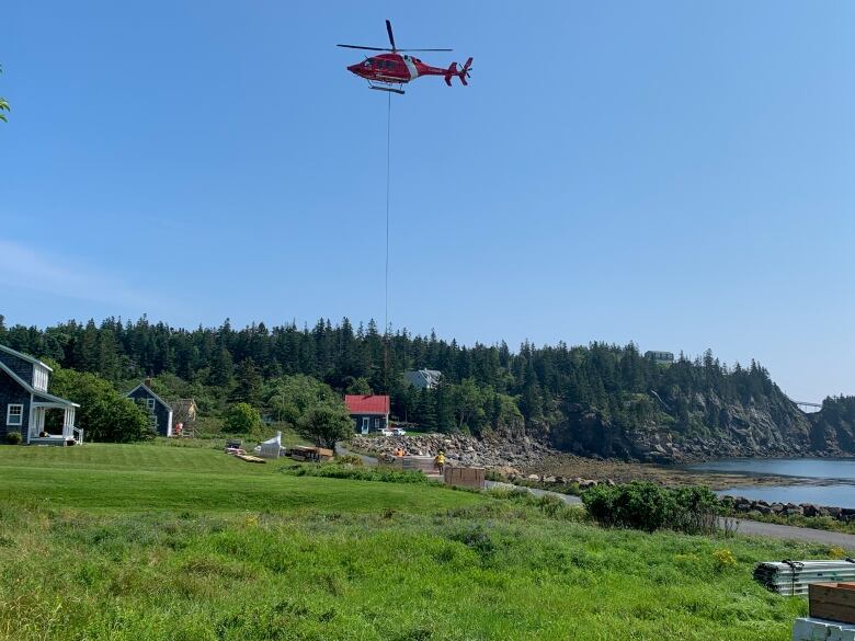 A red helicopter with a long rope carries wood to the shore of an island filled with dark green pine trees and several houses. 