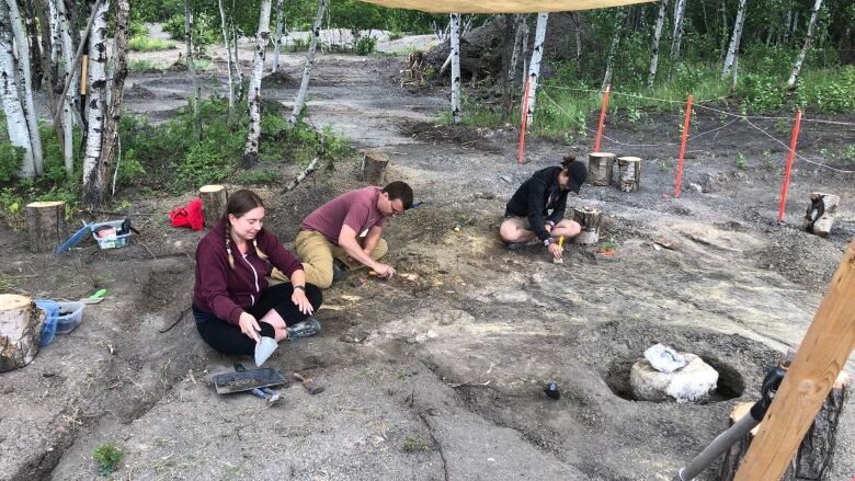 Three people sit on the ground under a tent and use tools to extract fossils.