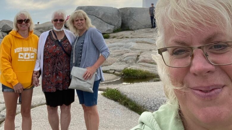 Three smiling women with blond hair stand on grey rocks with a lighthouse in the background, while another woman with blond hair and glasses stands to the right of them, smiling at the camera.