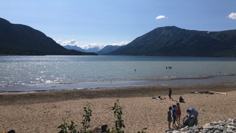 A few people are seen on the beach and in the water at a big lake with mountains in the distance.