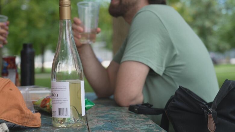 A bottle of wine is shown on a picnic table with a bearded man in the background holding a glass.