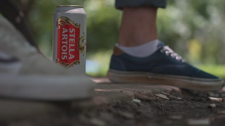 A can of Stella Artois is shown next to a man's show in the park.