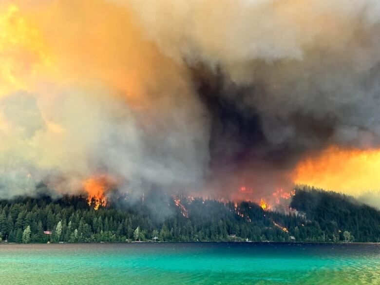 Flames rise from a forested hilltop next to a picturesque lake.