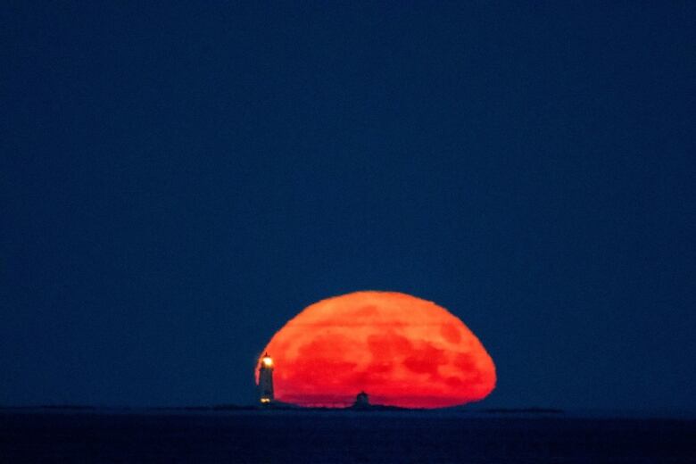 Full moon seen near the horizon and starting to rise in the sky in front of a lighthouse.