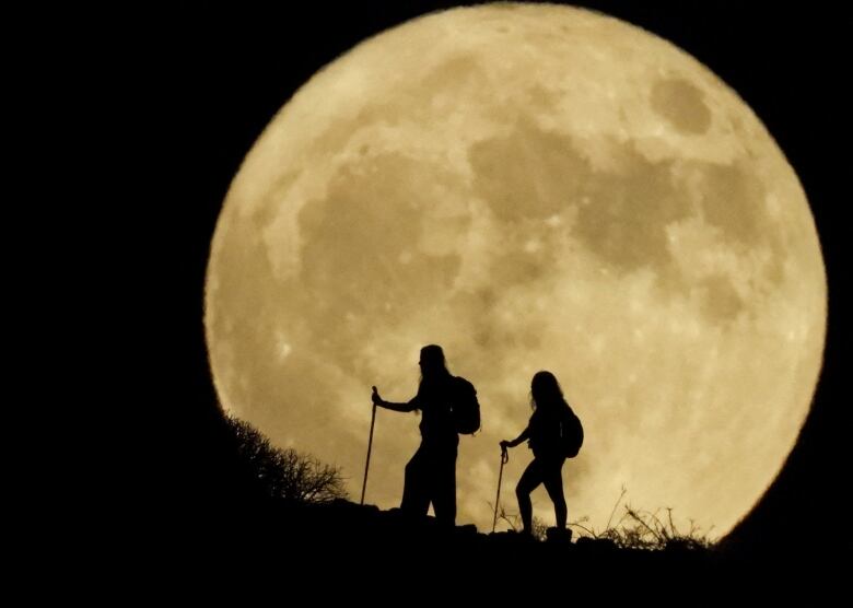 Silhouette of two hikers in front of a full moon.