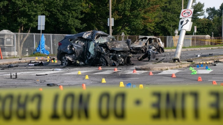 Behind a line of police tape, two cars can be seen crumpled and burned. 