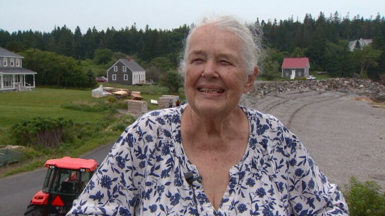 A woman in a blue and white floral pattern blouse smiles. In the background, there's a dirt road, a few houses, and lush green grass with dark green pine trees. 