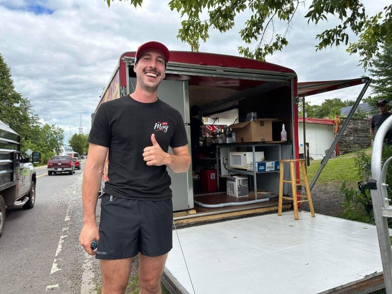 A man gives a thumbs up and stands in front of a trailer. 