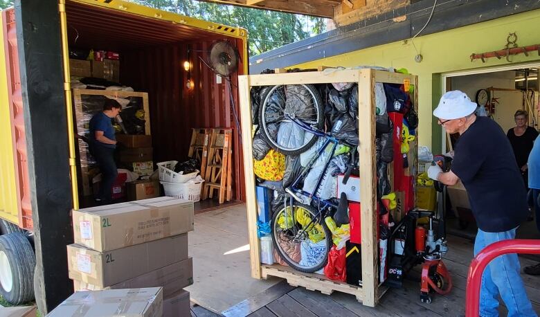 A man pushes a dolly with a box of donations, of which you can see a bike and other colorful objects, into the back of a container ship.