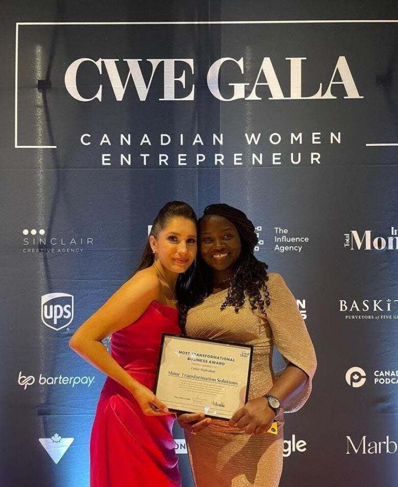 Two women standing in front of an event banner holding an award frame. 