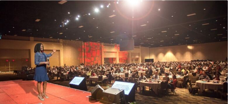 A conference room filled with people sitting and a woman standing on the podium addressing the people. 