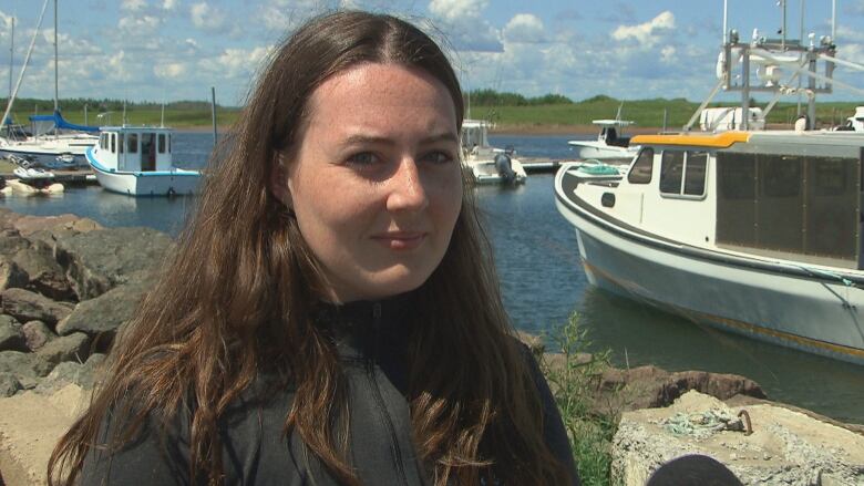 A woman with long brown hair standing in front of a marina with fishing boats in it.