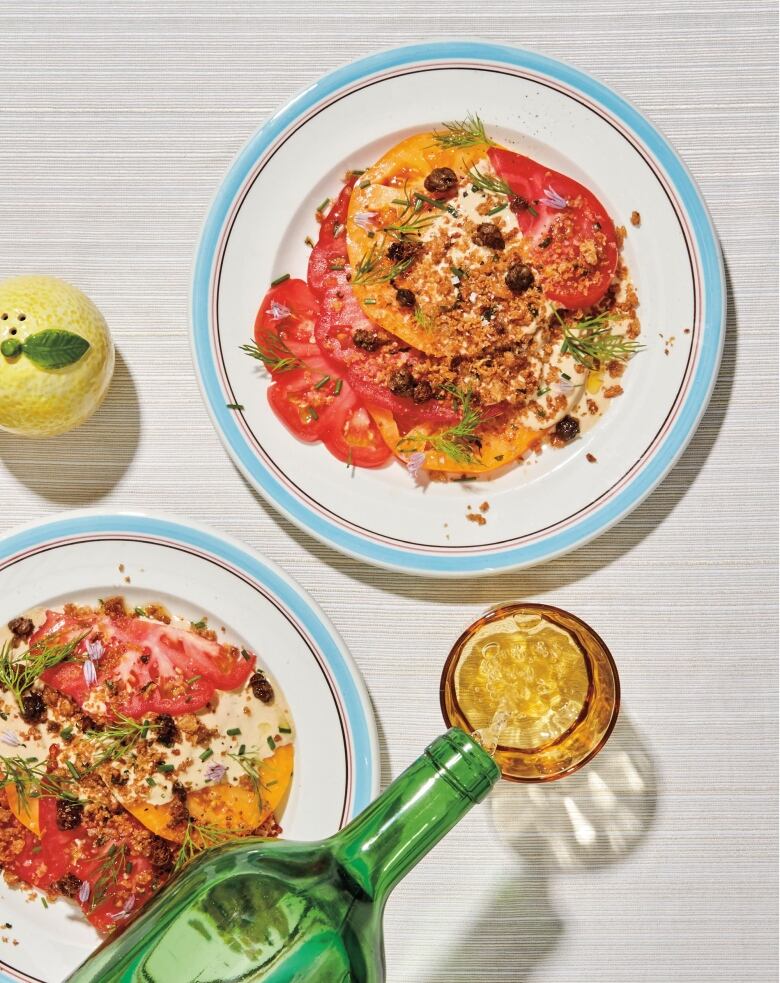 overhead shot of 2 blue and white plates with slices of tomatoes on them, topped with a creamy sauce, breadcrumbs, fried capers and fresh dill. They're on a grey table and a green wine bottle is pouring white wine into a yellow glass beside the plates. 