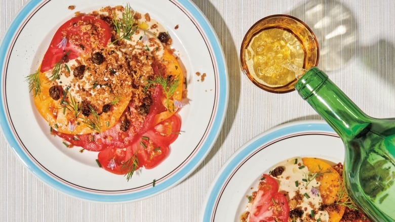 overhead shot of 2 blue and white plates with slices of tomatoes on them, topped with a creamy sauce, breadcrumbs, fried capers and fresh dill. They're on a grey table and a green wine bottle is pouring white wine into a yellow glass beside the plates.