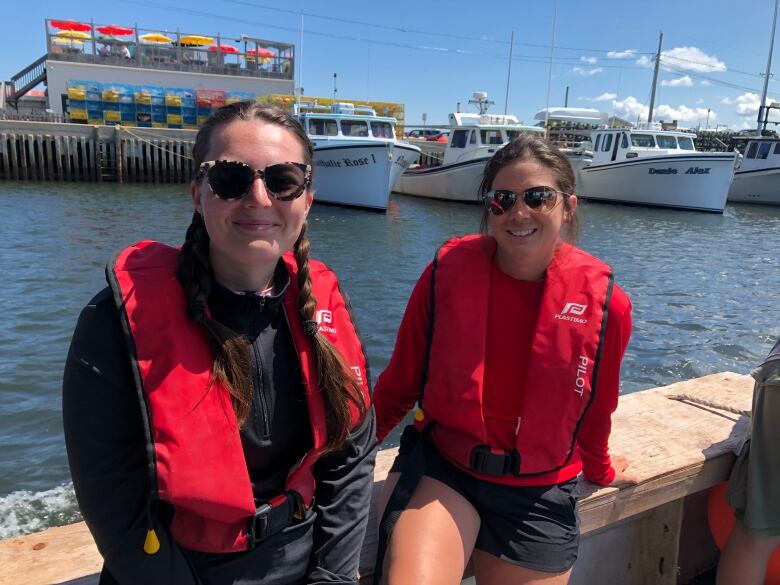 Two women in red life jackets sit on the wharf in sunglasses with the wharf behind them.