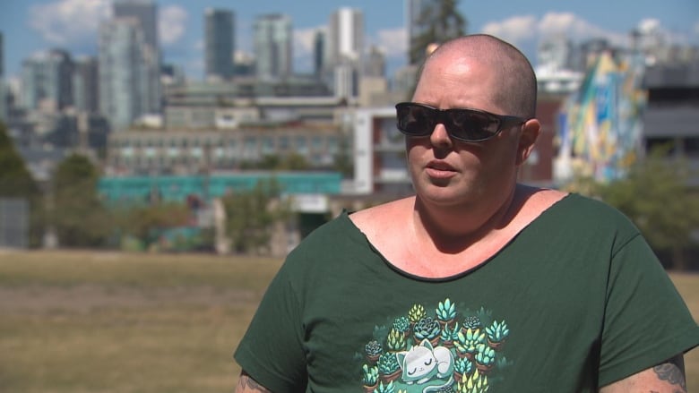 A woman with a shaved head wearing sunglasses and a green T-shirt with a circular floral emblem speaks framed by downtown Vancouver's highrises in the background.