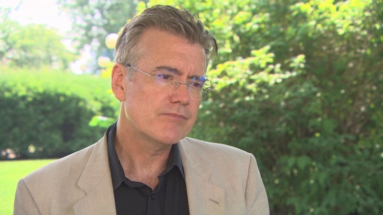 A man in a black shirt and tan jacket speaks to a reporter off camera.