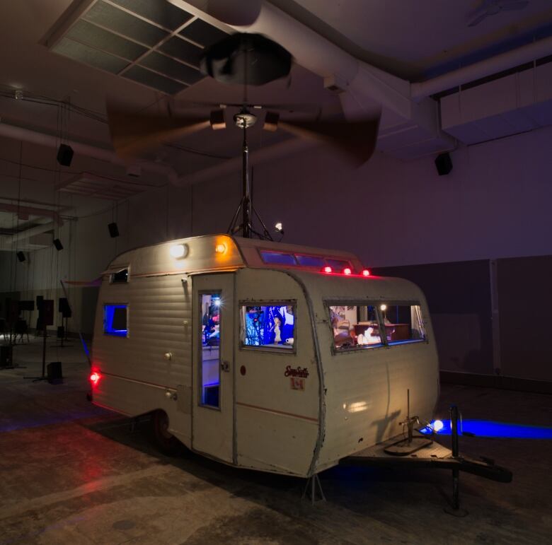 A small vintage camping trailer, lit with blue light from within, appears inside a darkened warehouse.