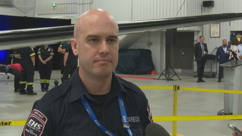 A bald white man is seen in a paramedic uniform, speaking to reporters in an airplane hangar.