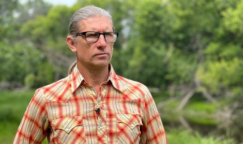 A man in a plaid shirt stands in front of trees.