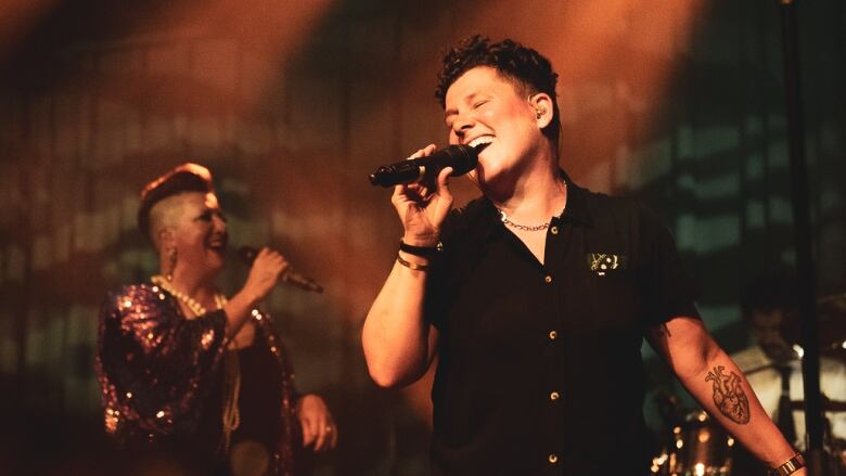 A person with short brown hair is singing into a mic. They have a short sleeved shirt on and a heart shaped tattoo on their outstretched forearm.  In the background is a person wearing pearls and a sequined gown also singing into a microphone. 