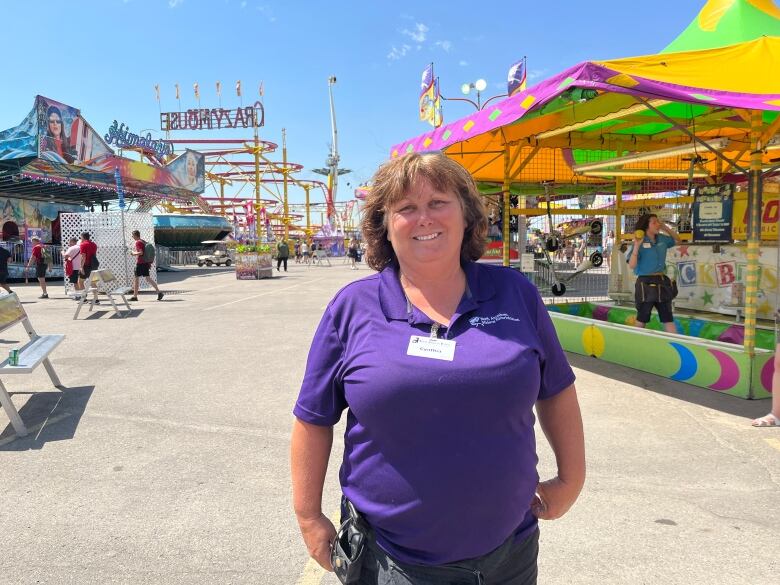 A woman in a purple branded t-shirt with carnival games and rides behind her