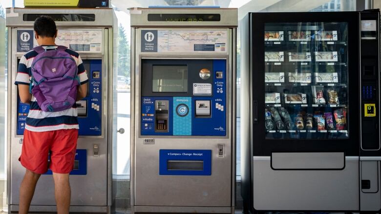 A person stands in front of a ticket vending machine at a SkyTrain station. To the right, there is another ticket vending machine and a vending machine featuring meals like sushi.